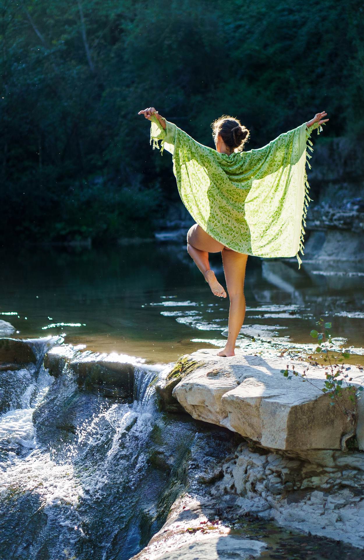 Stefania in equilibrio su un piede sulla roccia sopra la cascata, kimono verde aperto nella luce
