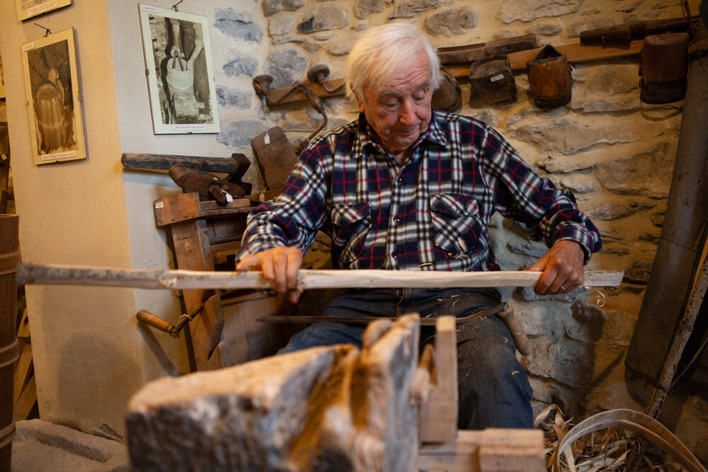 Il maestro bigonaio intaglia una doga di legno con la roncola, seduto nella bottega tra attrezzi antichi